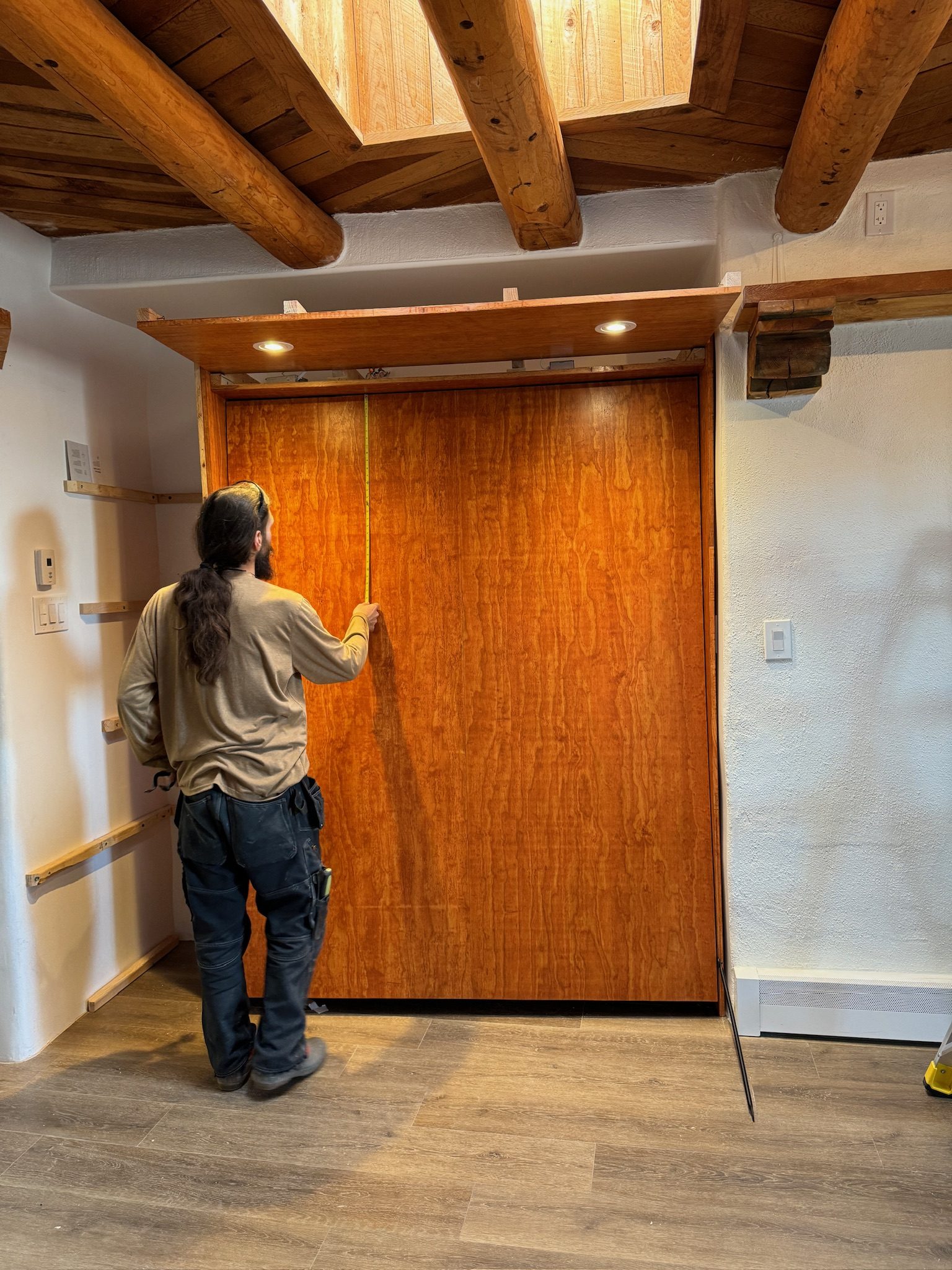 A carpenter running a tape down the full height of the closed cherry face, checking the fit against the opening.