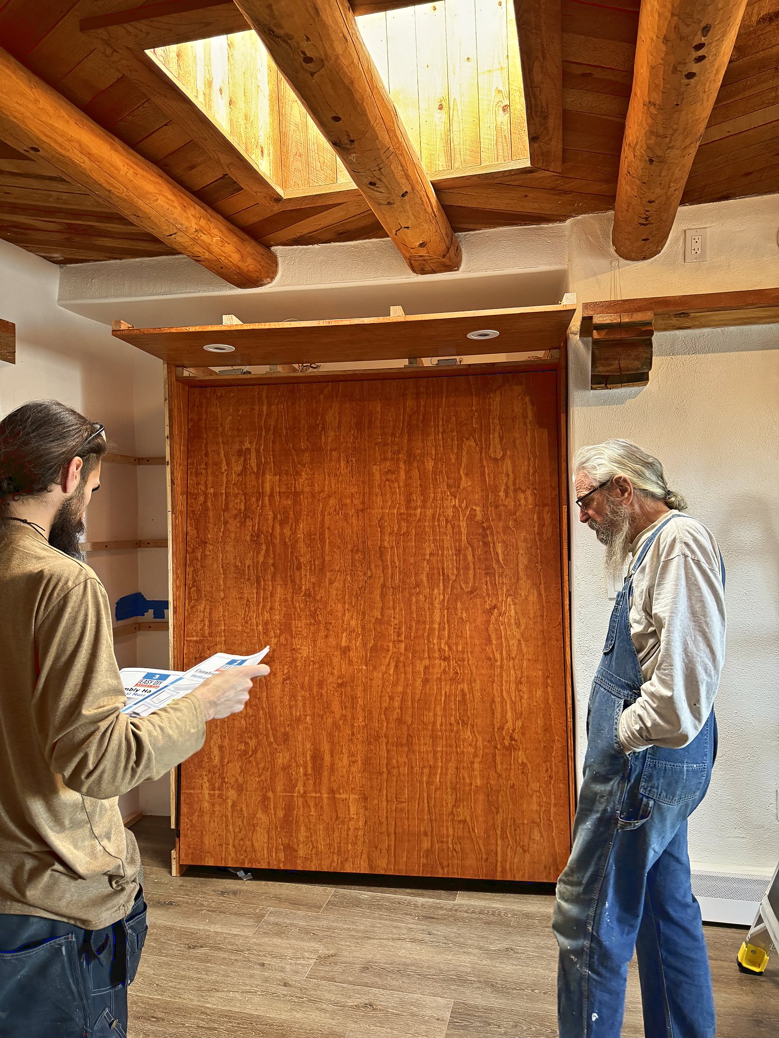 Two carpenters either side of the finished cherry cabinet, one reading the fitting instructions and the older man in denim overalls looking it over.