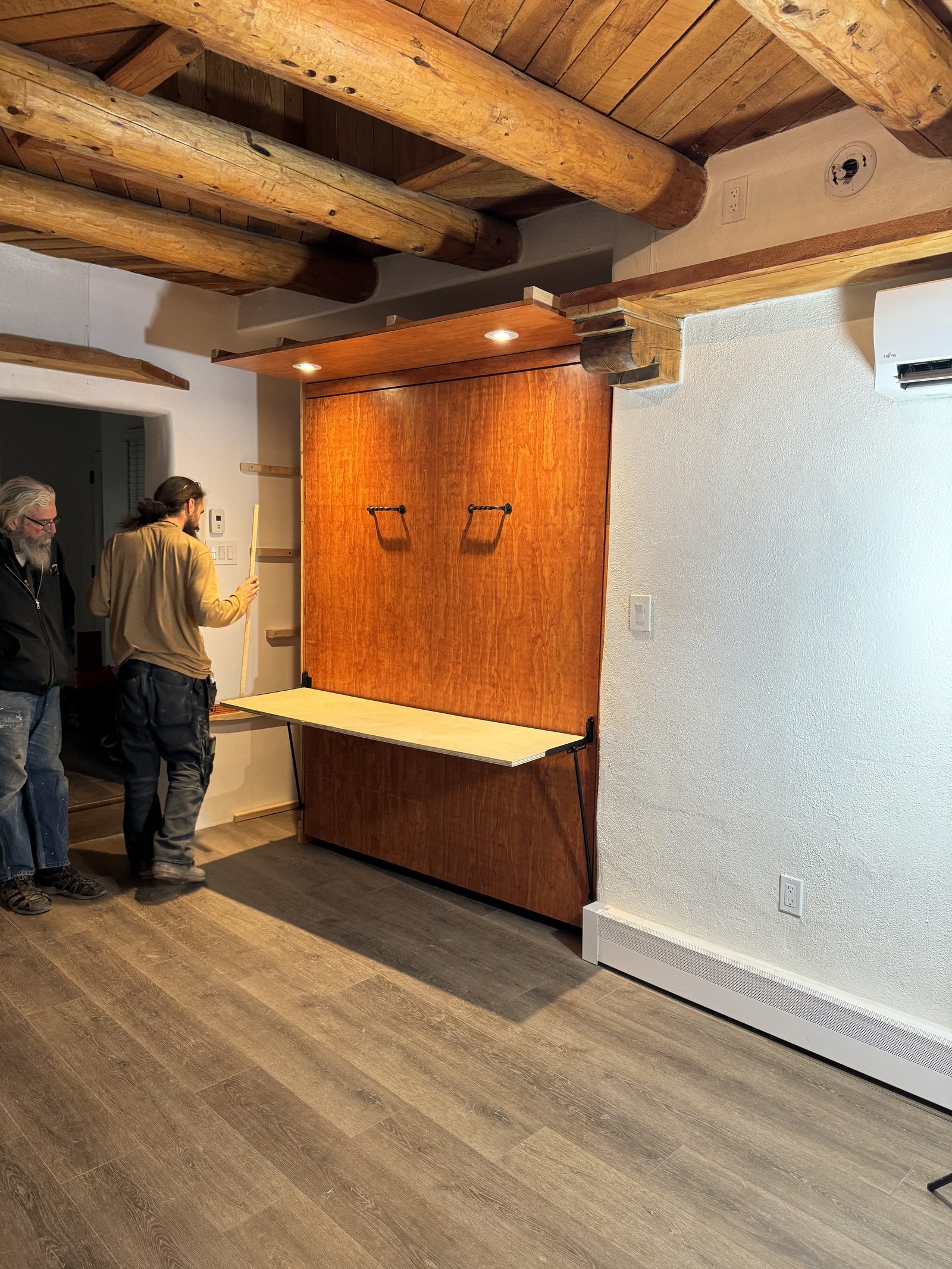 The desk top being fitted: the bed shut away, the worktop out on its brackets, and two men measuring along the shelves beside it.