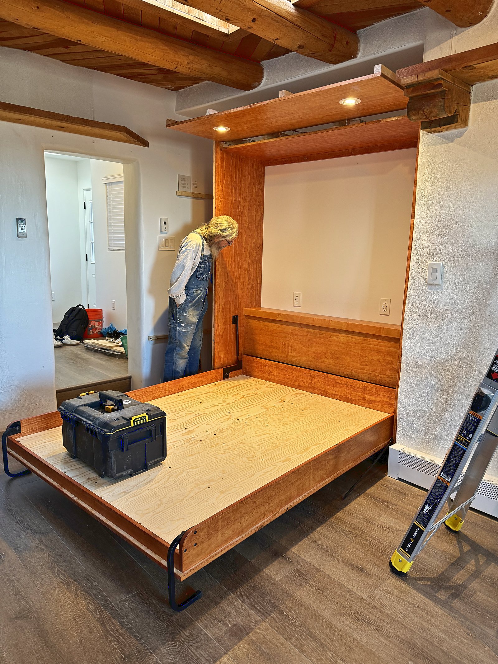 The bed folded down out of the cabinet for the first time, the bare platform out on its steel legs with a toolbox sitting on it, and the carpenter bent to the fixings behind.