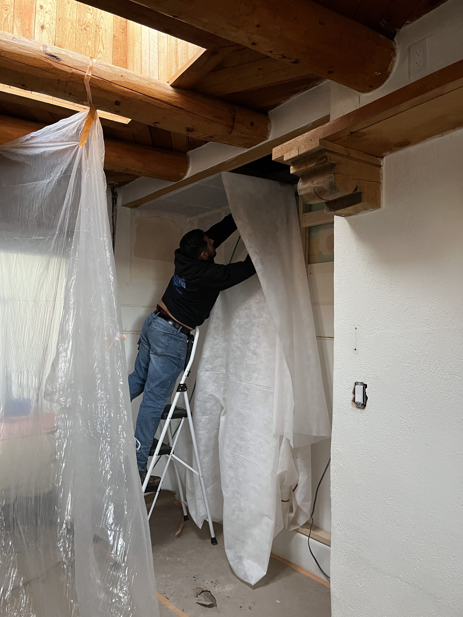 The alcove sheeted in plastic and paper with a carpenter up a stepladder fixing the covering to the vigas before the joinery goes in.