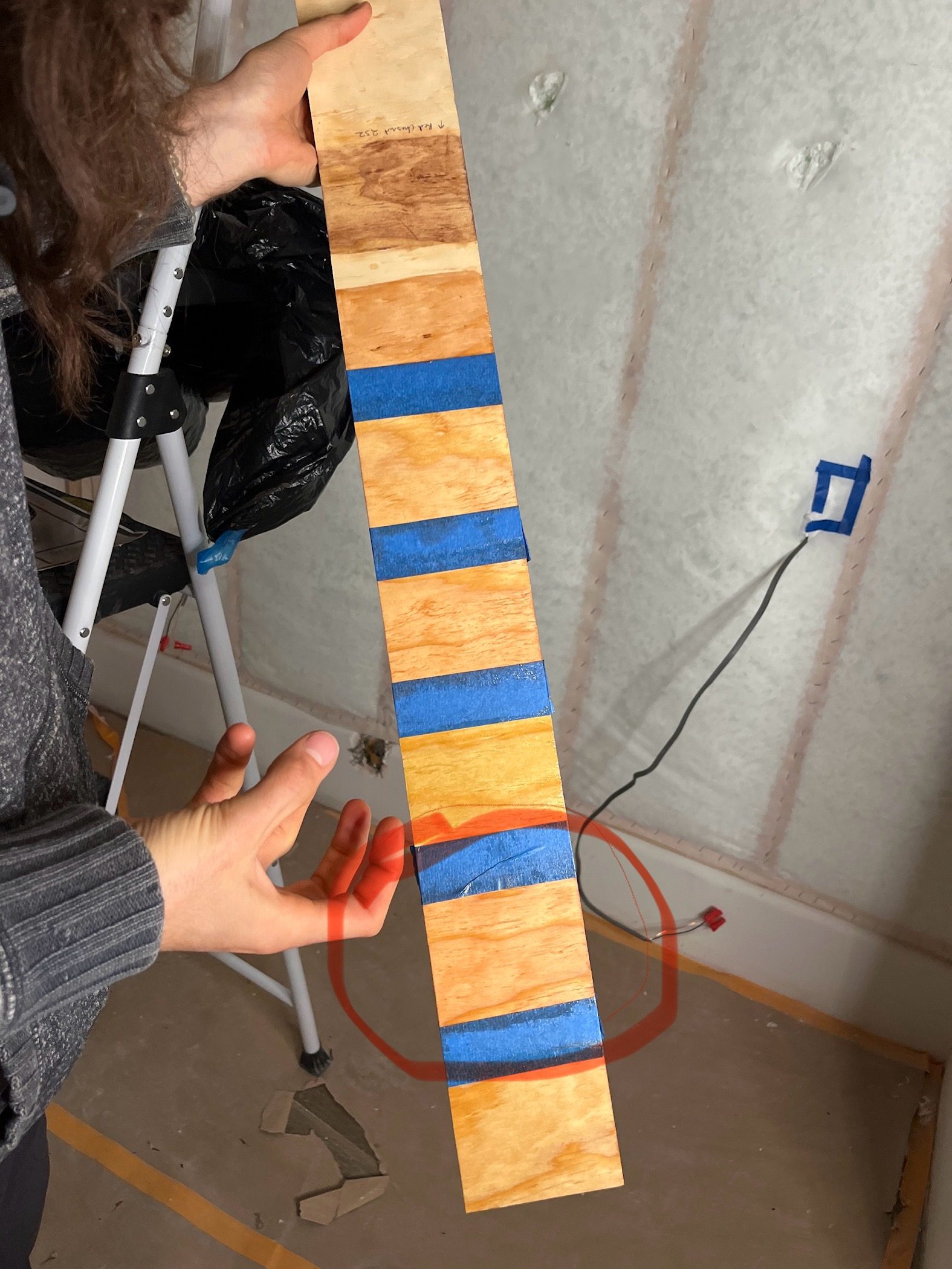 A long stain sample board held up against the wall, six finishes taped off along the plywood, with one circled.