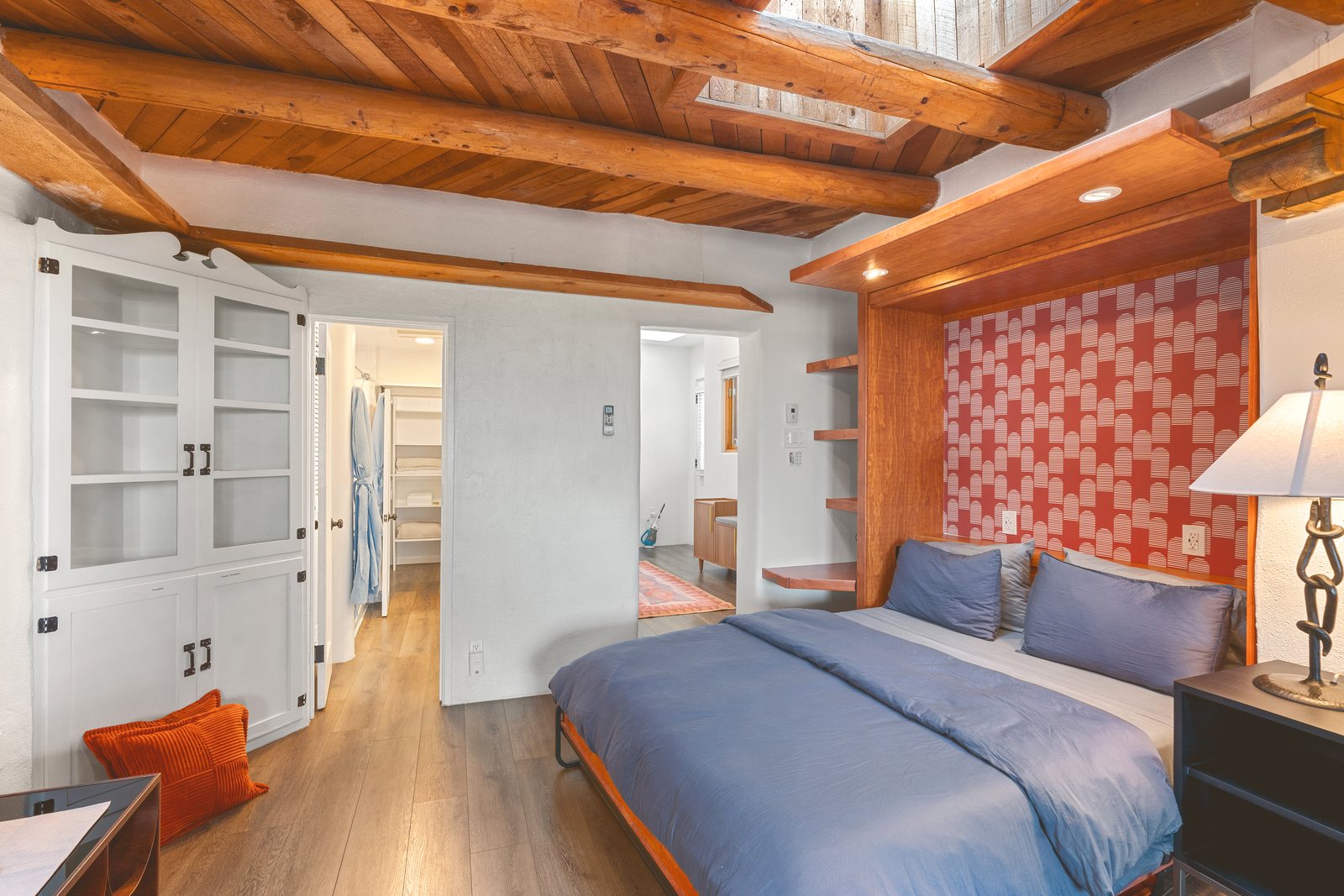 The bed down and made up: blue linens under the lit cherry head, the coral patterned wall behind, vigas and a skylight overhead and the walk-in closet standing open beyond.