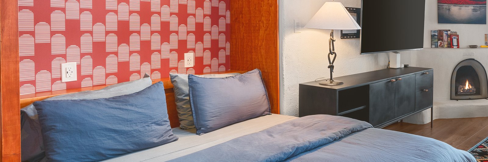 The casita made up for the night: the queen bed dressed in soft blue against the red patterned tile wall, a lamp lit beside it, vigas and a wood plank ceiling overhead, and the kiva fireplace around the corner.