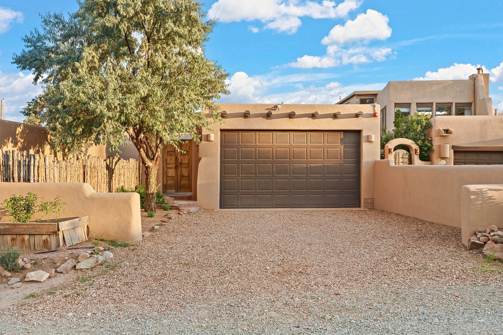 The front of the compound on a clear day: a tan adobe wall and garage under a mature tree, a carved timber gate through to the courtyard on the left, and a bright sky with scattered cloud above.