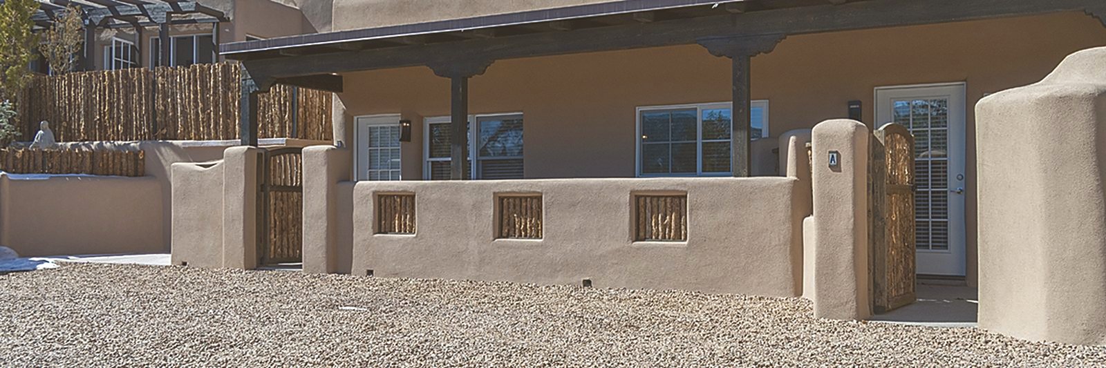 The long covered portal running the length of the compound: stuccoed adobe walls, carved wooden posts and beams overhead, coyote-pole gates opening to each unit's private entrance, gravel underfoot and deep blue sky above.