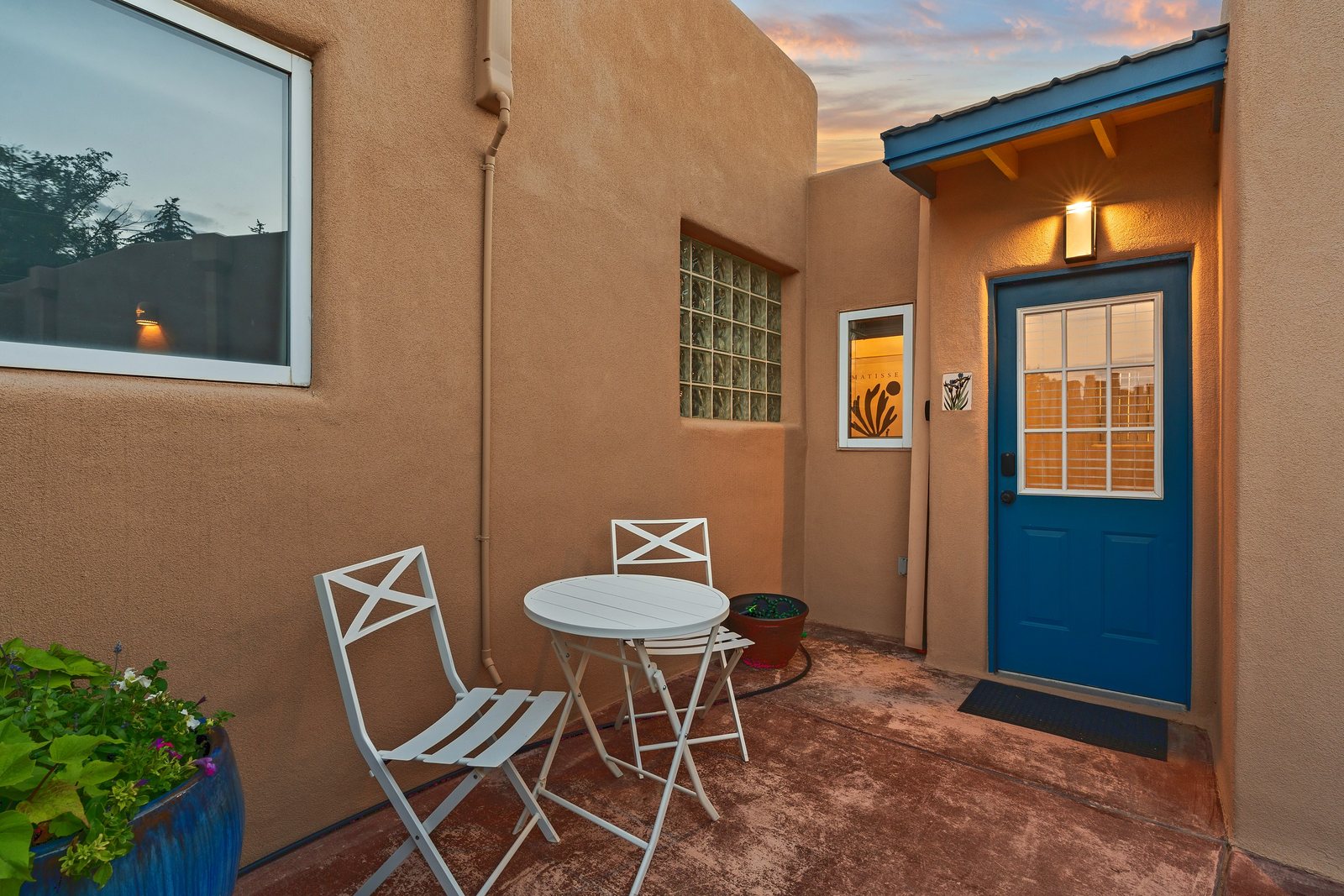 The private courtyard at dusk: a teal front door lit warm under its own lamp, a white bistro table and chairs against the adobe wall, and a planter of trailing greenery in the foreground.