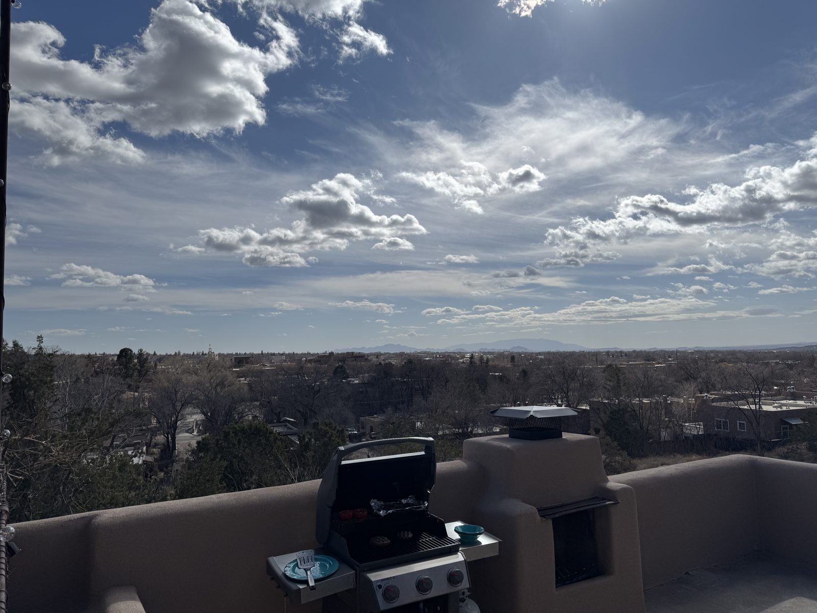 The deck on a bright day: the grill and the outdoor kiva fireplace along the parapet wall, the town spread out below, and a wide sky of scattered cloud above.