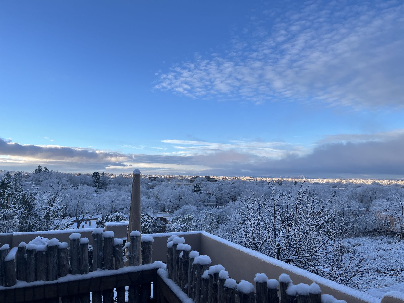 The deck under fresh snow: every post of the coyote-pole fence capped white, the parapet wall edged in snow, and the valley beyond turned pale to the horizon.