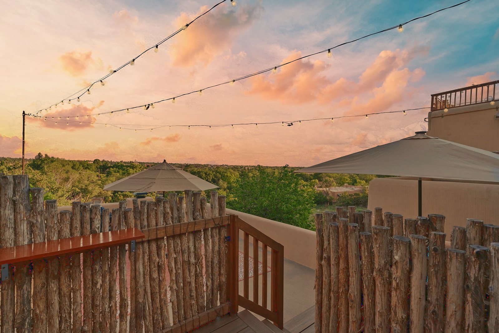 Sunset from the deck: pink cloud across a wide sky, strings of bulbs overhead, the coyote-pole gate and rail in the foreground, and the tree canopy running out to the horizon.