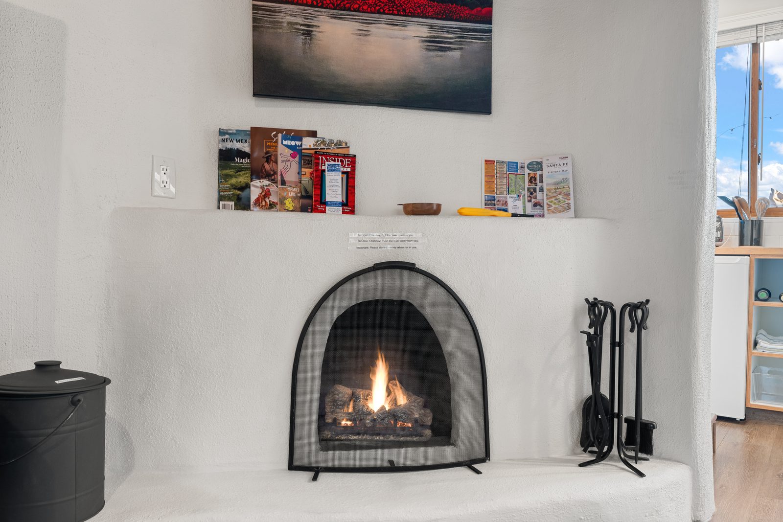 The hand-plastered kiva fireplace with a fire lit behind its mesh screen, the curved white hearth built into the corner, an ash bucket to one side and wrought-iron fire tools to the other.