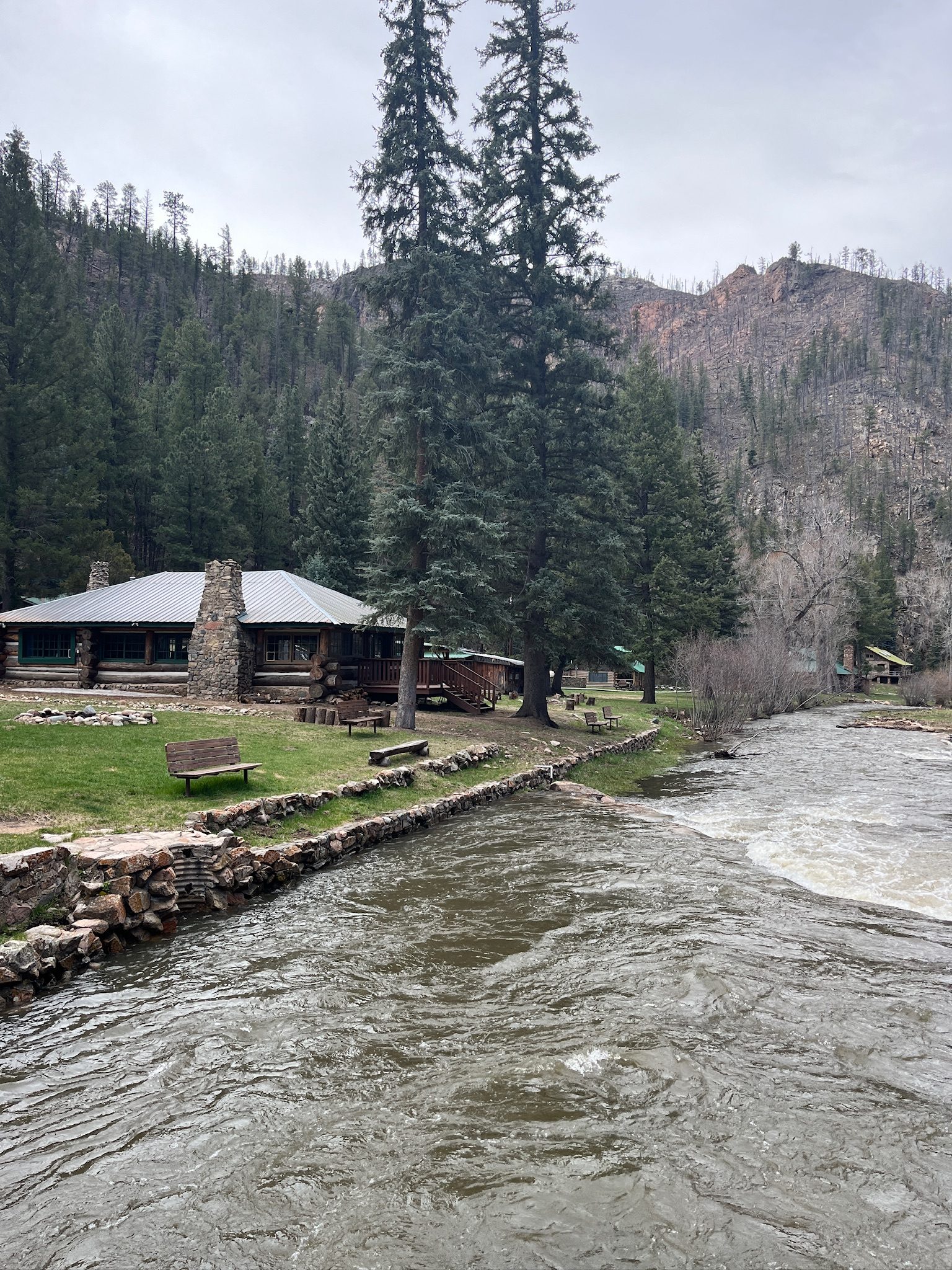 The Pecos running high past a log lodge with a stone chimney, spruce towering over the bank.