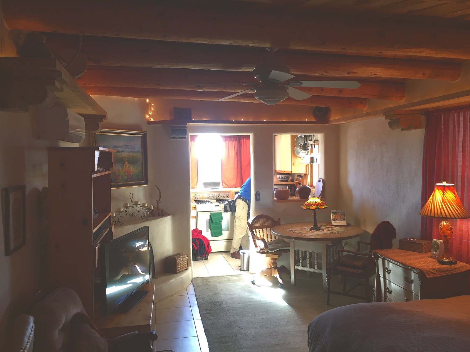 Inside the casita in 2018: the same viga ceiling overhead, a ceiling fan, heavy dark furniture, a round table and the kitchen through the opening.