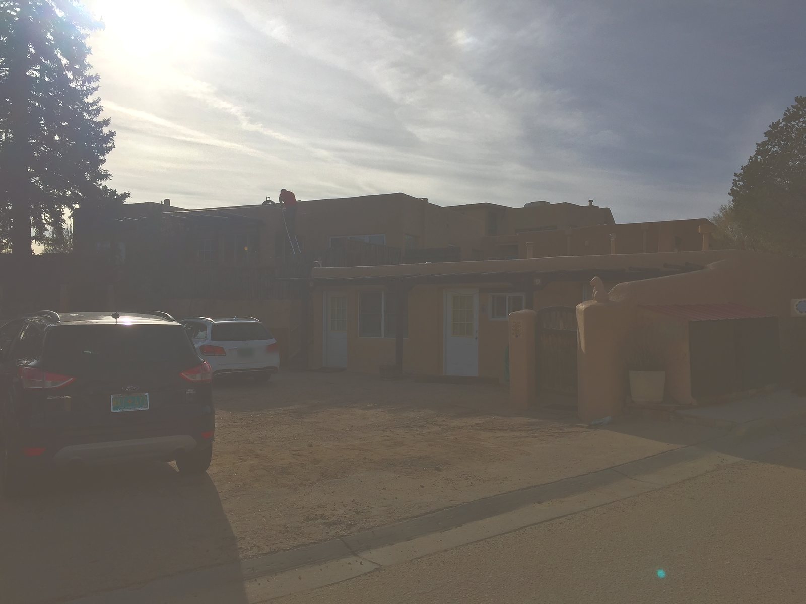 The compound from the street on the 2018 visit, low winter sun behind it: the long adobe wall, the carved gate, a corrugated porch roof, and a roofer up a ladder on the far unit.