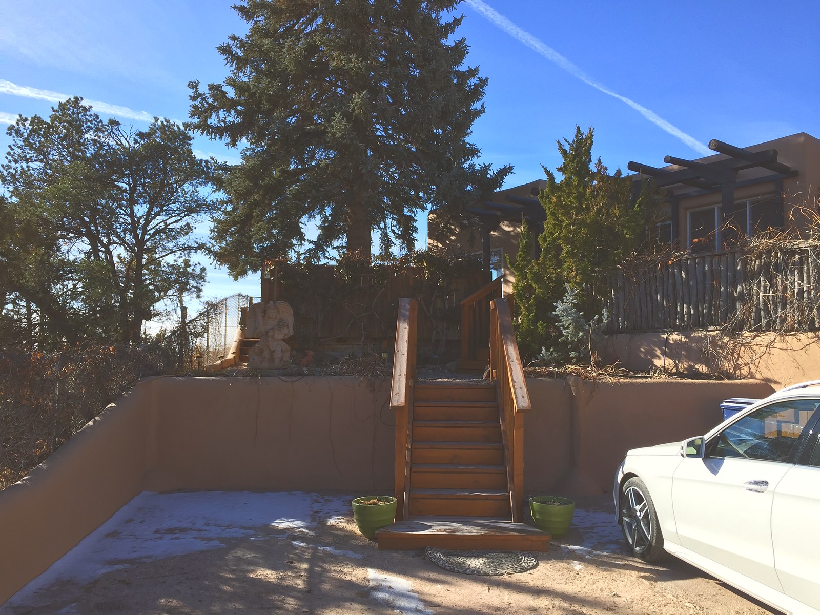 The gravel court in 2018: a bare timber stair climbing to the units, a low plastered wall, the big spruce behind it and a car parked on the gravel.