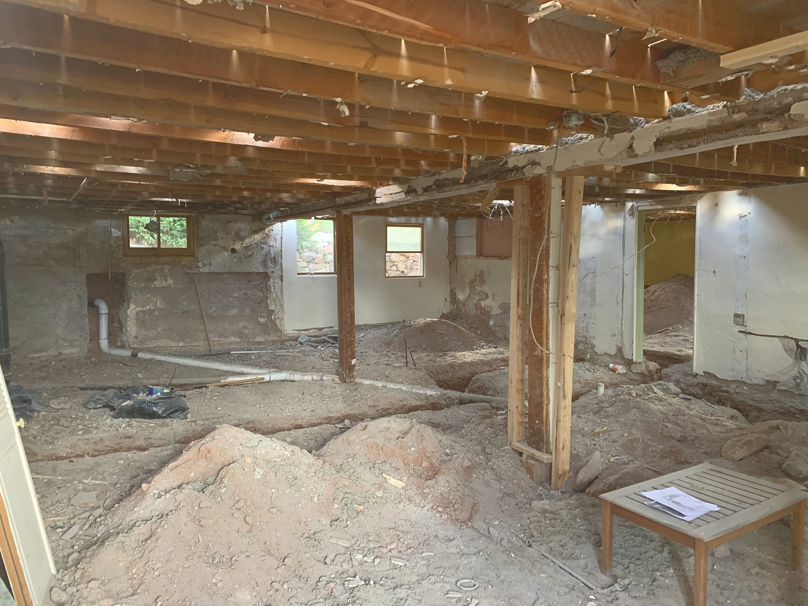 June 2020: a unit taken back to the dirt, ceiling joists open above, temporary posts holding the span, and the old floor dug out for new plumbing.