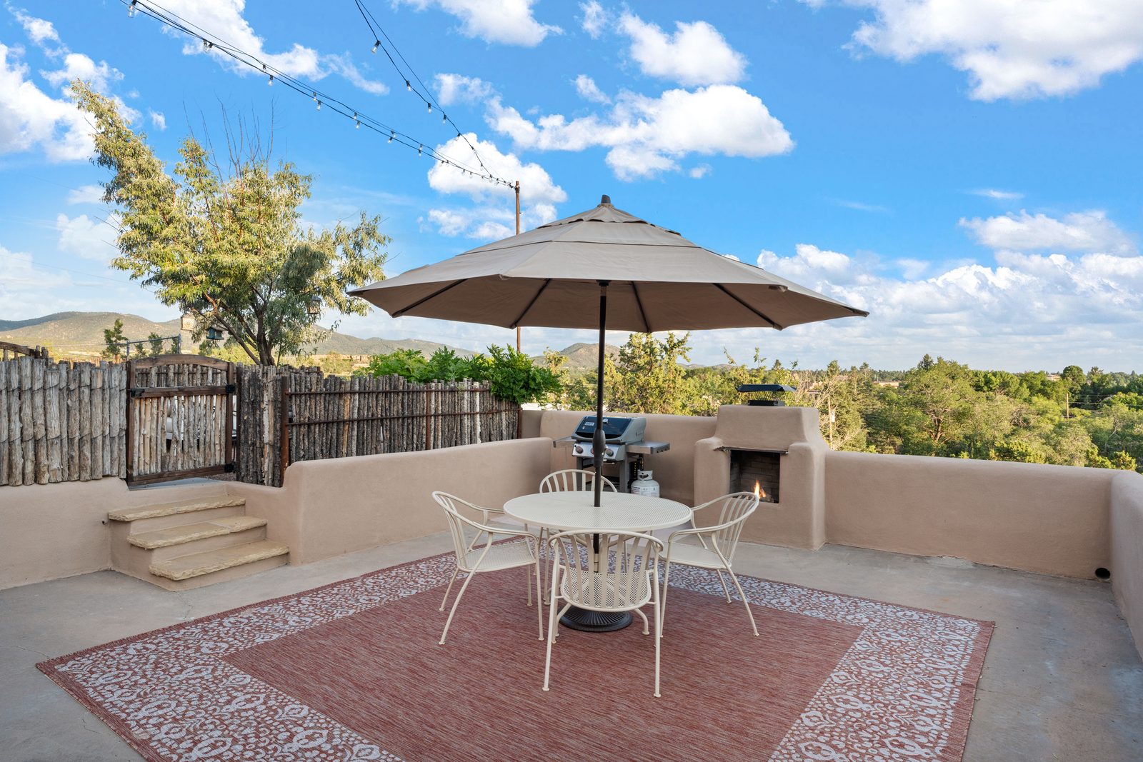 The rooftop deck in the early evening: a canvas umbrella over a table and chairs on a patterned outdoor rug, a gas grill and a stuccoed outdoor kiva fireplace along the parapet, string lights overhead, and the foothills beyond the coyote-pole fence.