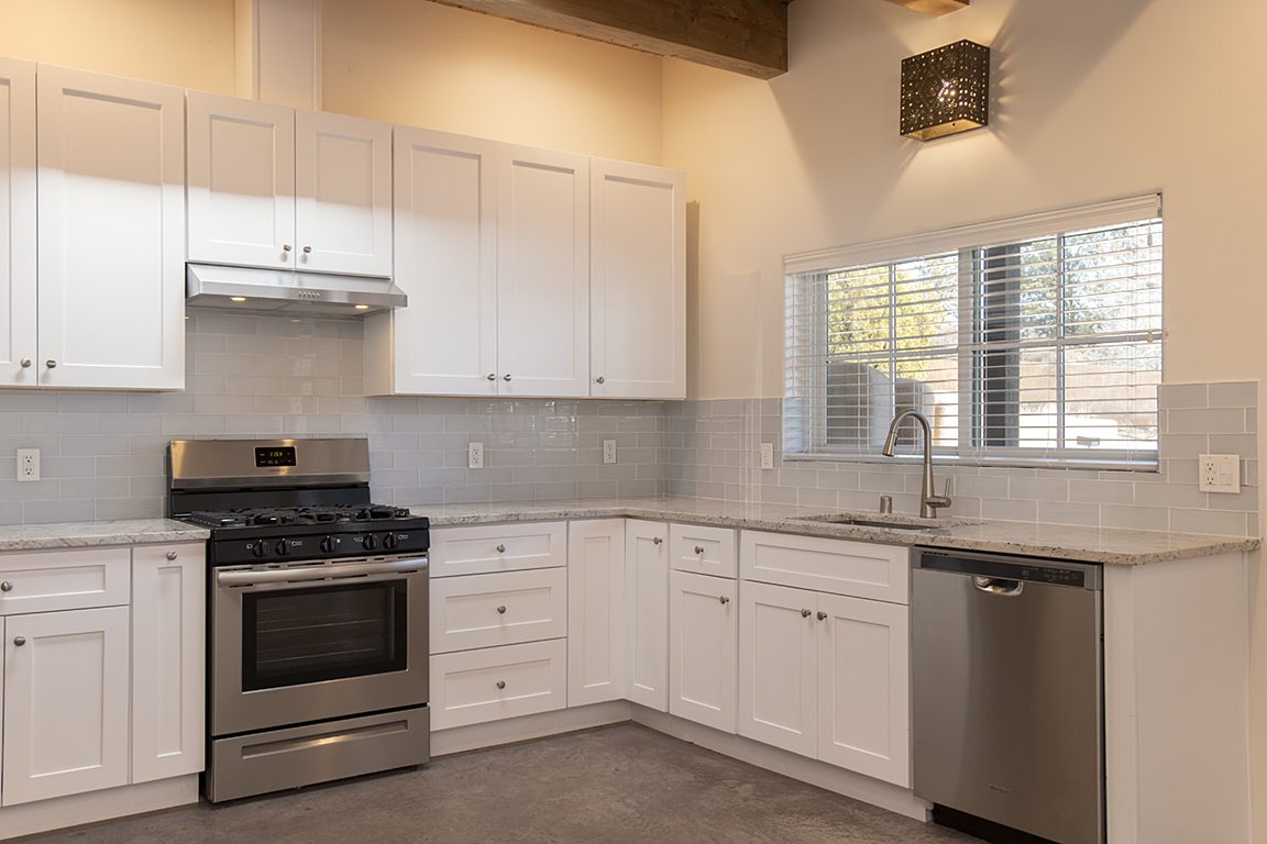 Another side of Unit B's kitchen: white shaker cabinets, a gas range under the hood, the dishwasher beside the sink and a punched-tin light overhead.