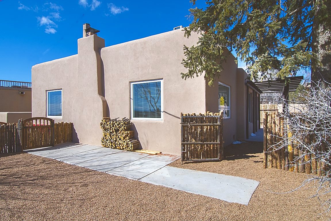 Unit C from the side yard: smooth adobe walls and a corner chimney under a deep blue sky, split firewood stacked against the wall and coyote-pole gates on both sides.