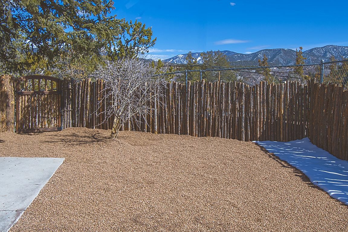 Unit C's gravelled yard behind its coyote-pole fence, with the Sangre de Cristos rising over the fence line and a patch of snow in the shade.