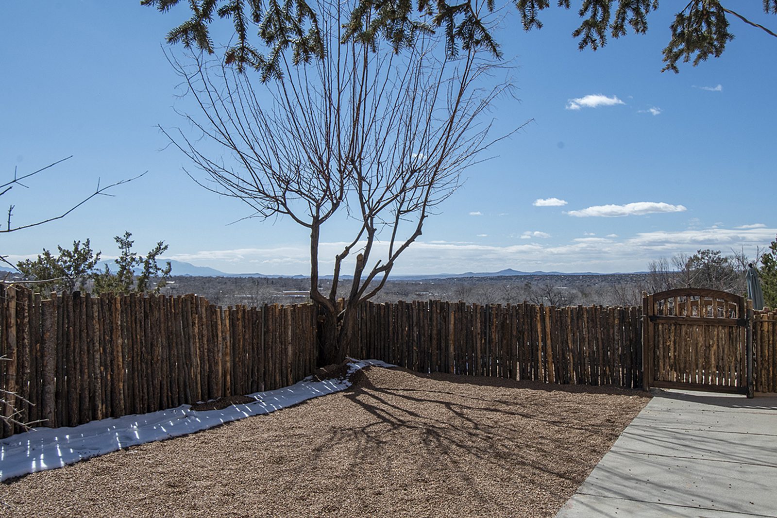 Unit C's gated yard: gravel, a mature tree, coyote-pole fencing, and an open view to the mountains beyond.