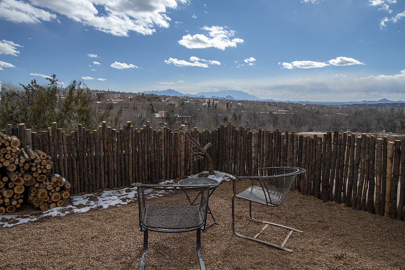 Unit D's rebuilt yard: new coyote-pole fence around a gravel court, two iron chairs and a stacked woodpile, with all of Santa Fe and the mountains beyond the fence line.