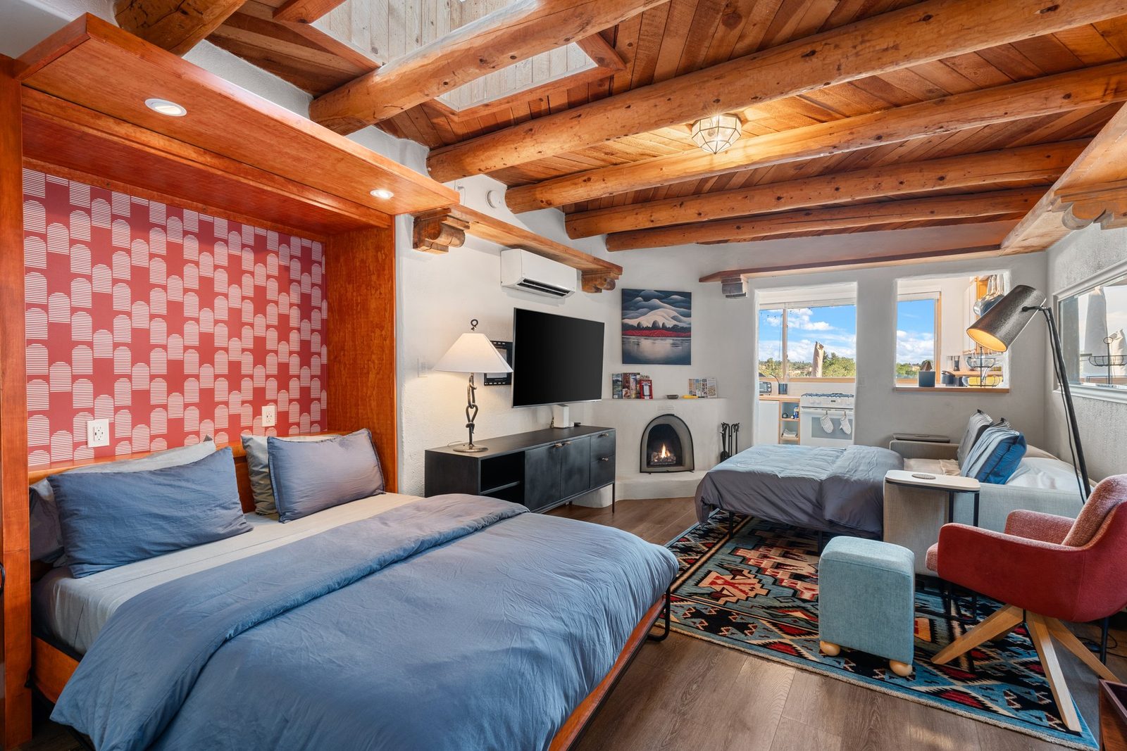 The queen bed made up in blue against a red patterned tile wall, wood vigas overhead, the room open to the living space.