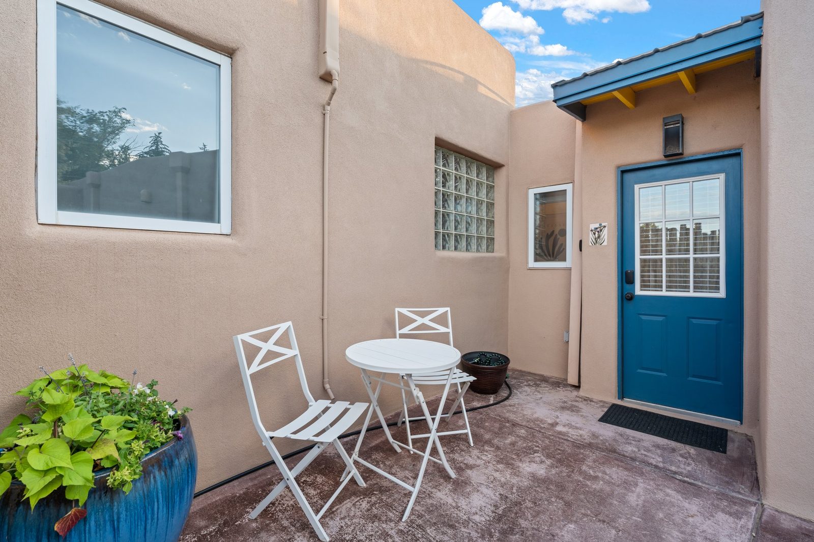The same corner today: the walled courtyard with its blue-painted door, glass-block window, potted plants and a small bistro table.