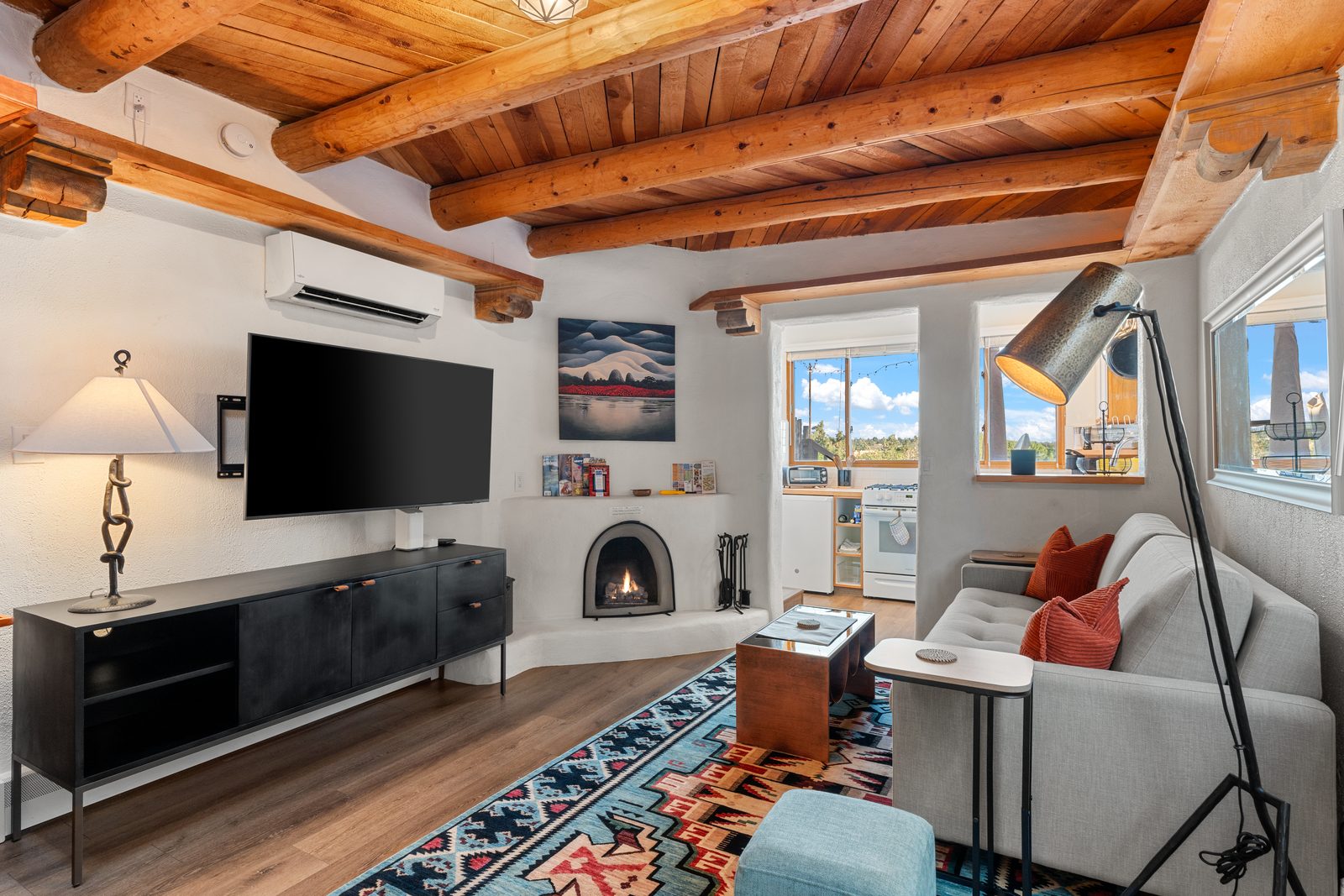 The casita's main room with a fire lit in the white plaster kiva fireplace, exposed viga ceiling, sofa, wall-mounted television, and a patterned wool rug on wood floors.