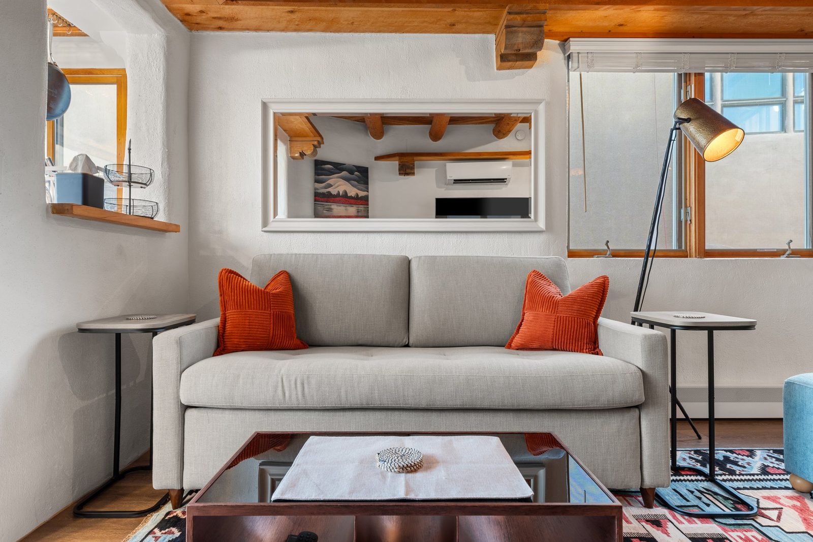 The casita's main room: a pale sofa with rust cushions under framed desert prints, a wood-beamed ceiling, and the kiva fireplace around the corner.