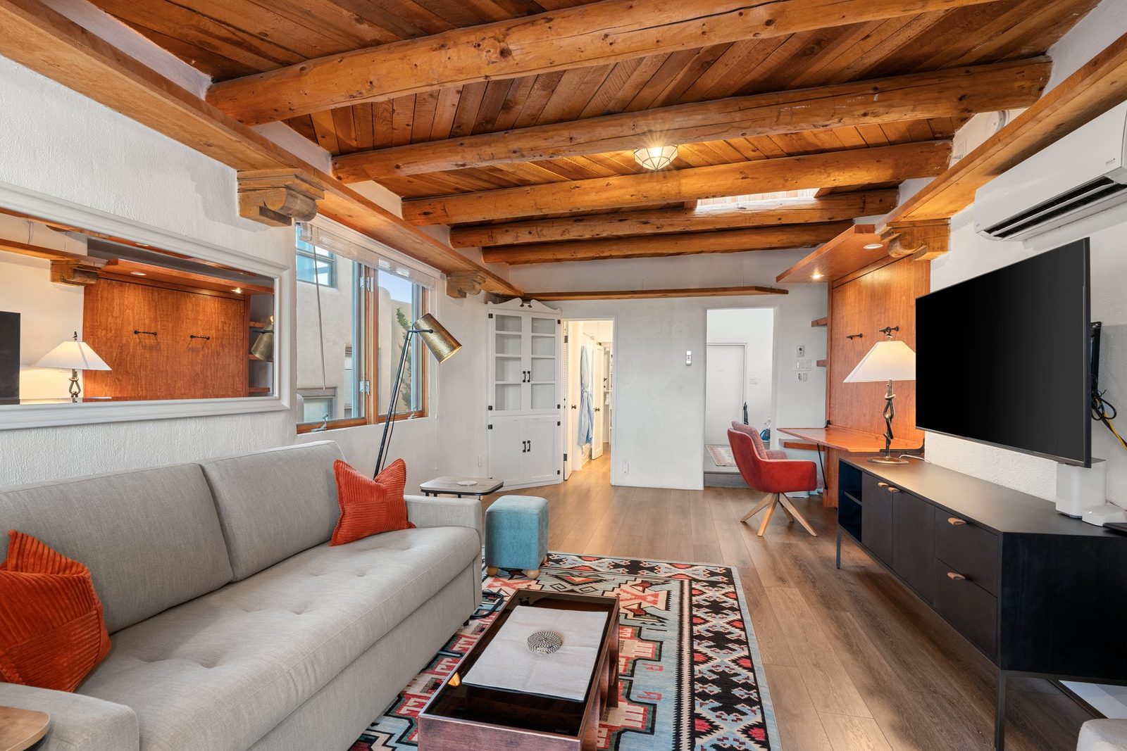 The living area with the television on the wall, a floor lamp, patterned wool rug over wood floors, and windows on two sides.