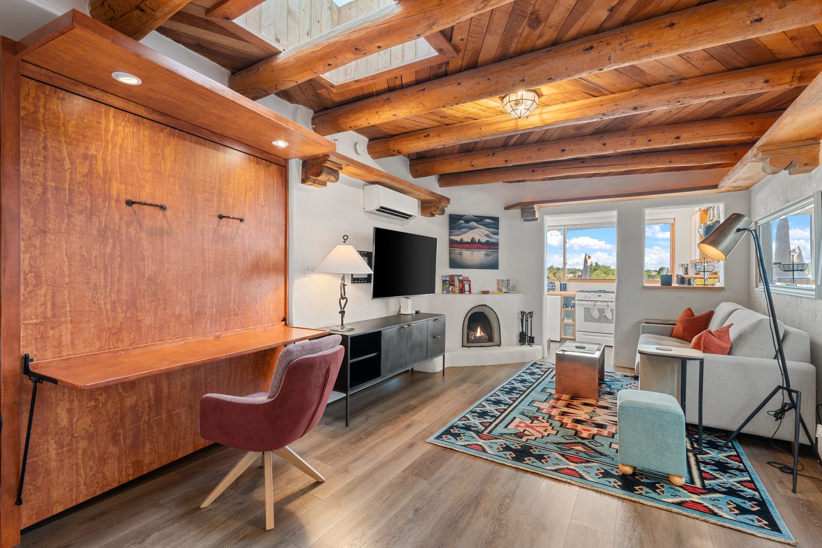 The same room now: the vigas cleaned back to the wood, a lit kiva fireplace, a wide desk that folds down from the cabinetry, a wool rug and the kitchen through the same opening.