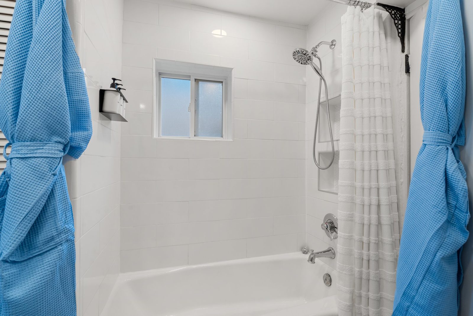 The full bathtub with a shower over it, blue towels and robes hung alongside a window.